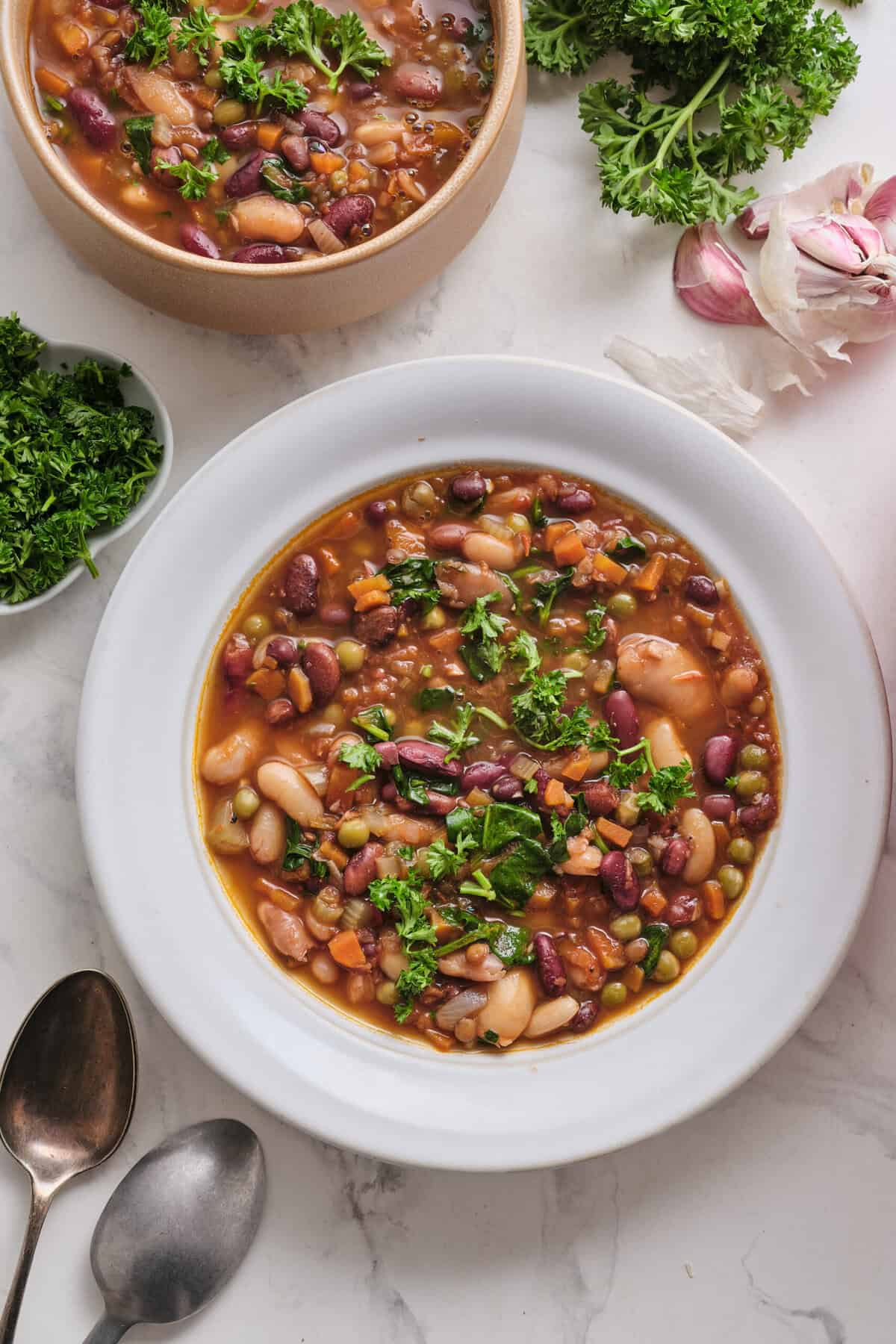 Hearty vegan bean vegetable soup with kidney beans, carrots, and spinach, garnished with fresh parsley, in a white bowl on a white marble surface.