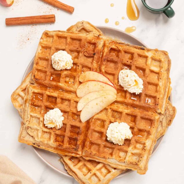 Golden Belgian waffles topped with whipped cream and fresh apple slices, served with cinnamon sticks and syrup on a white marble background. Perfect for healthy breakfast ideas and nutritious meal planning.
