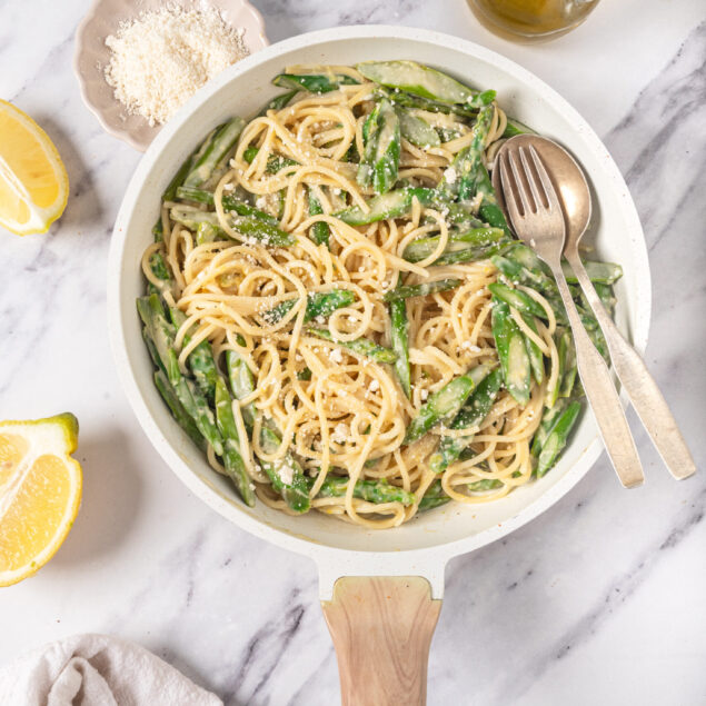 Creamy lemon garlic pasta with asparagus in a white skillet, topped with grated Parmesan cheese, on marble surface with lemon wedges, olive oil, and tools for healthy meal recipes.