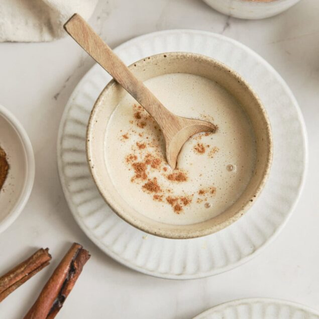 Creamy cinnamon spiced almond milk latte in a rustic ceramic cup on a white surface, surrounded by cinnamon sticks, perfect for healthy breakfast recipes and nutritious beverages.