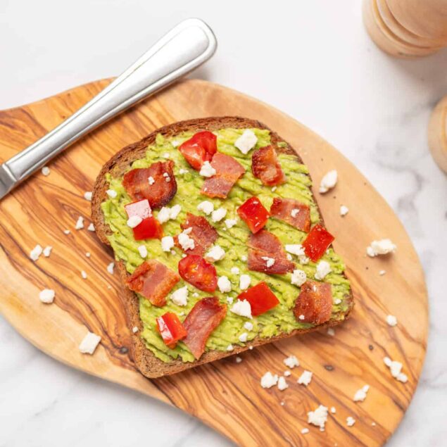 Creamy avocado toast topped with crispy bacon, cherry tomatoes, and crumbled cheese on whole grain bread, served on a wooden cutting board.