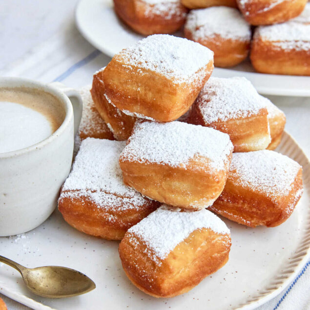 Fluffy, golden-brown fried donuts topped with powdered sugar, served on a white plate with a cup of coffee, perfect for breakfast or brunch.