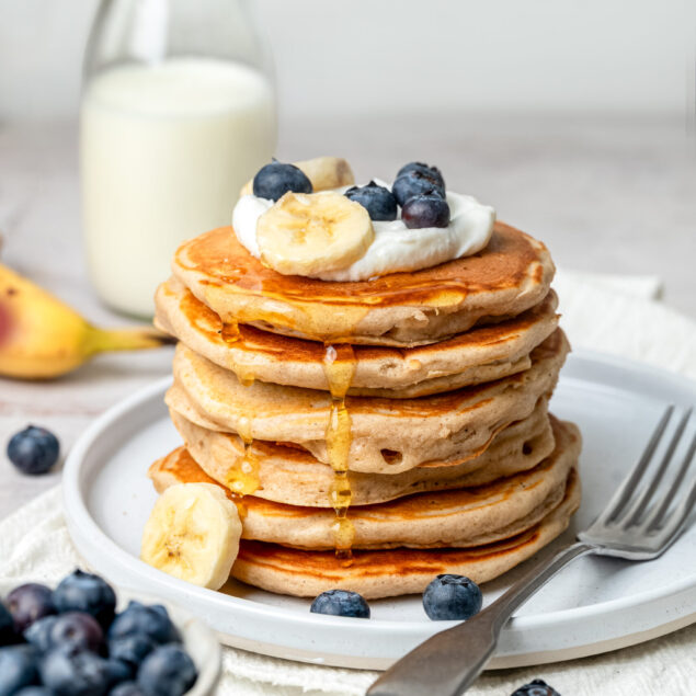 Fluffy homemade pancake stack topped with fresh bananas, blueberries, and whipped cream, served with maple syrup and a glass of milk, perfect for healthy breakfast recipes.