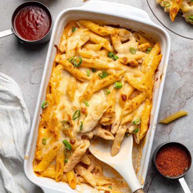 Creamy baked penne pasta with melted cheese, chicken, and green onions in a white casserole dish on a light surface.