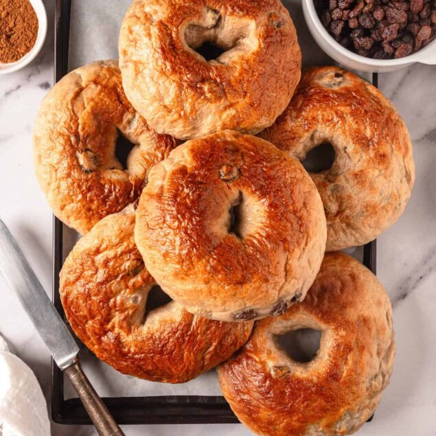 Fluffy homemade bagels with a golden-brown crust on a black tray, served with dried raisins, ground cinnamon, and a knife for spreading or slicing, perfect for healthy breakfast or snack options.