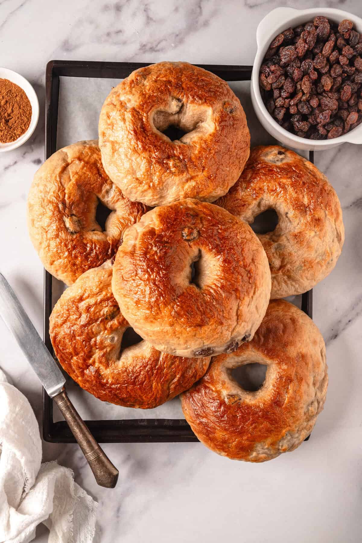 Fluffy homemade bagels with a golden-brown crust on a black tray, served with dried raisins, ground cinnamon, and a knife for spreading or slicing, perfect for healthy breakfast or snack options.