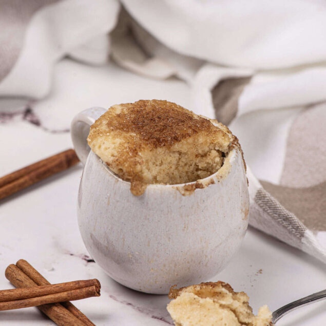 Creamy baked cinnamon mug cake topped with cinnamon sugar, served in a white mug on marble surface with cinnamon sticks and a spoonful of cake. Perfect healthy dessert or snack from Food Faith Fitness.
