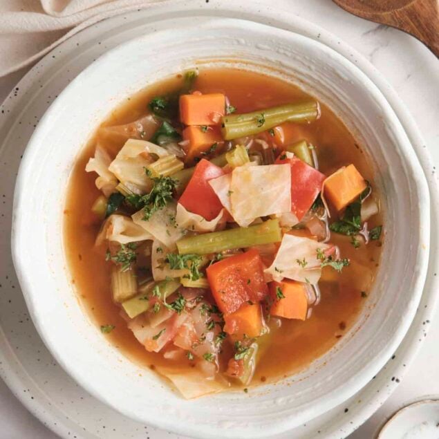 Vegan vegetable soup with fresh carrots, celery, cabbage, and tomatoes in a clear broth, served in a white bowl on a rustic background.