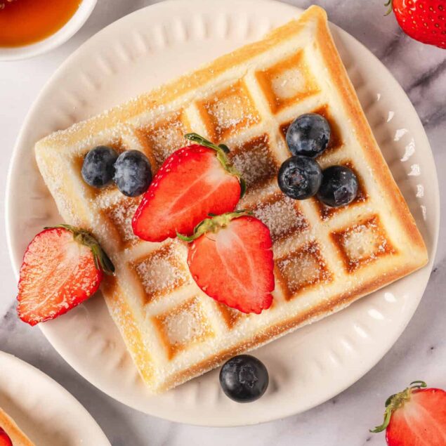 Crispy Belgian waffle topped with fresh strawberries blueberries and powdered sugar on white plate with honey and strawberries in background.