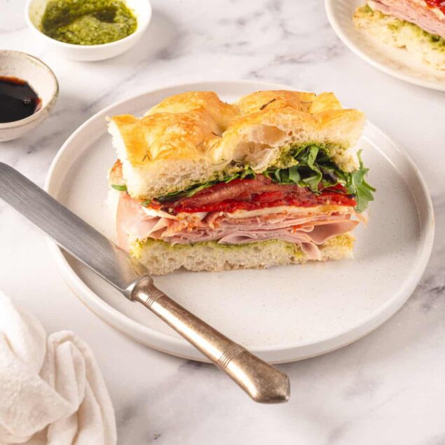 Juicy turkey and ham sandwich on a crusty baguette with fresh lettuce, tomato, and pesto side dip on marble surface.