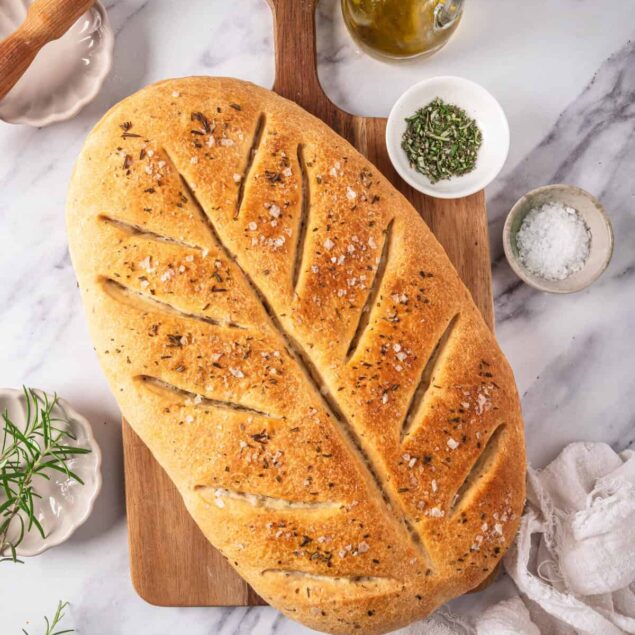 Focaccia bread on wooden cutting board with herbs, salt, olive oil, and baking tools, showcasing healthy, delicious homemade bread recipes from Food Faith Fitness.