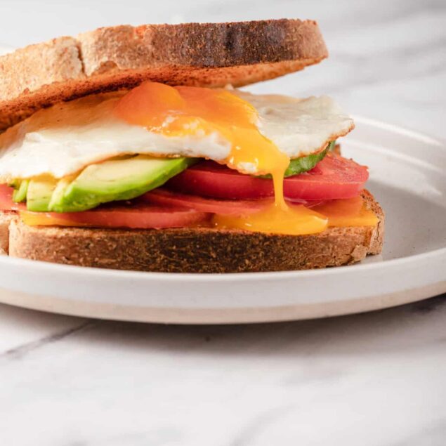 Soft-boiled egg breakfast sandwich with avocado, tomato, and whole grain bread on a white plate. Healthy, nutritious, and easy breakfast idea from Food Faith Fitness.