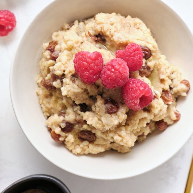 Creamy raspberry chocolate chip oatmeal served in a white bowl, topped with fresh raspberries and served with fresh blueberries and a small bowl of brown sugar on a white background.