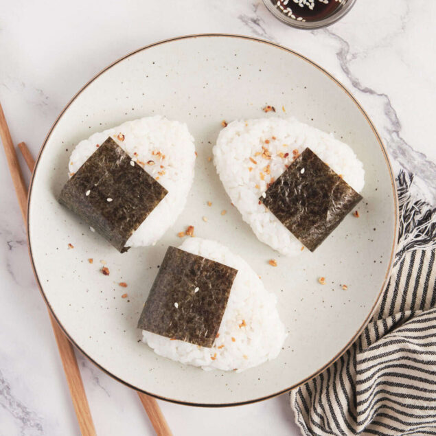 Fluffy Japanese onigiri rice balls with nori seaweed and sesame seeds on a stylish white plate, served with soy sauce and a side of toasted sesame seeds, perfect for healthy meal ideas.