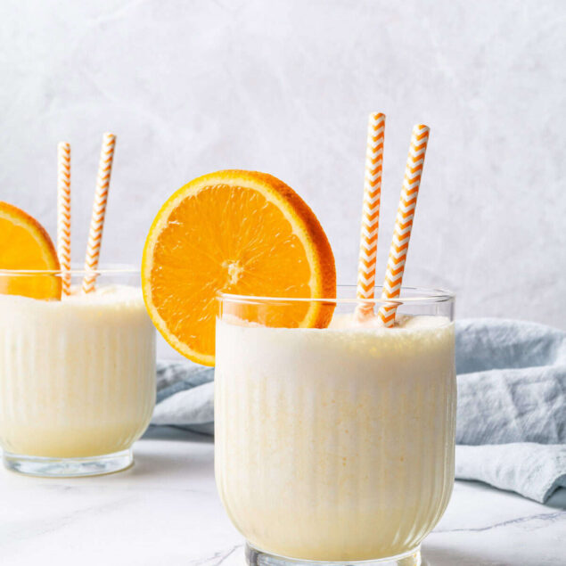 Creamy orange smoothie with fresh orange slices and striped paper straws on a white marble surface, perfect for health-conscious, nutritious recipes.