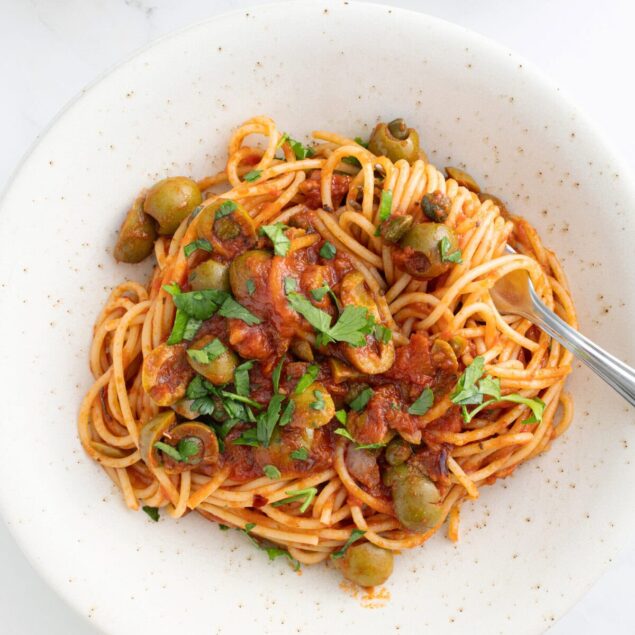 Creamy vegan pasta with green olives, tomato sauce, and fresh herbs on a white plate. Delicious plant-based meal perfect for healthy eating and fitness enthusiasts.
