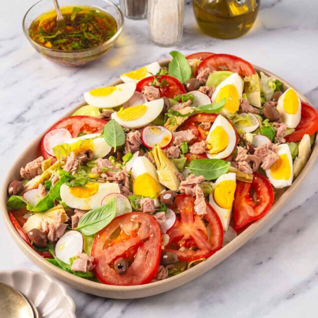 Fresh tuna niçoise salad with boiled eggs, tomatoes, radishes, artichokes, and basil on a white marble surface. Includes olive oil, seasonings, and vinaigrette for healthy, flavorful meal options.