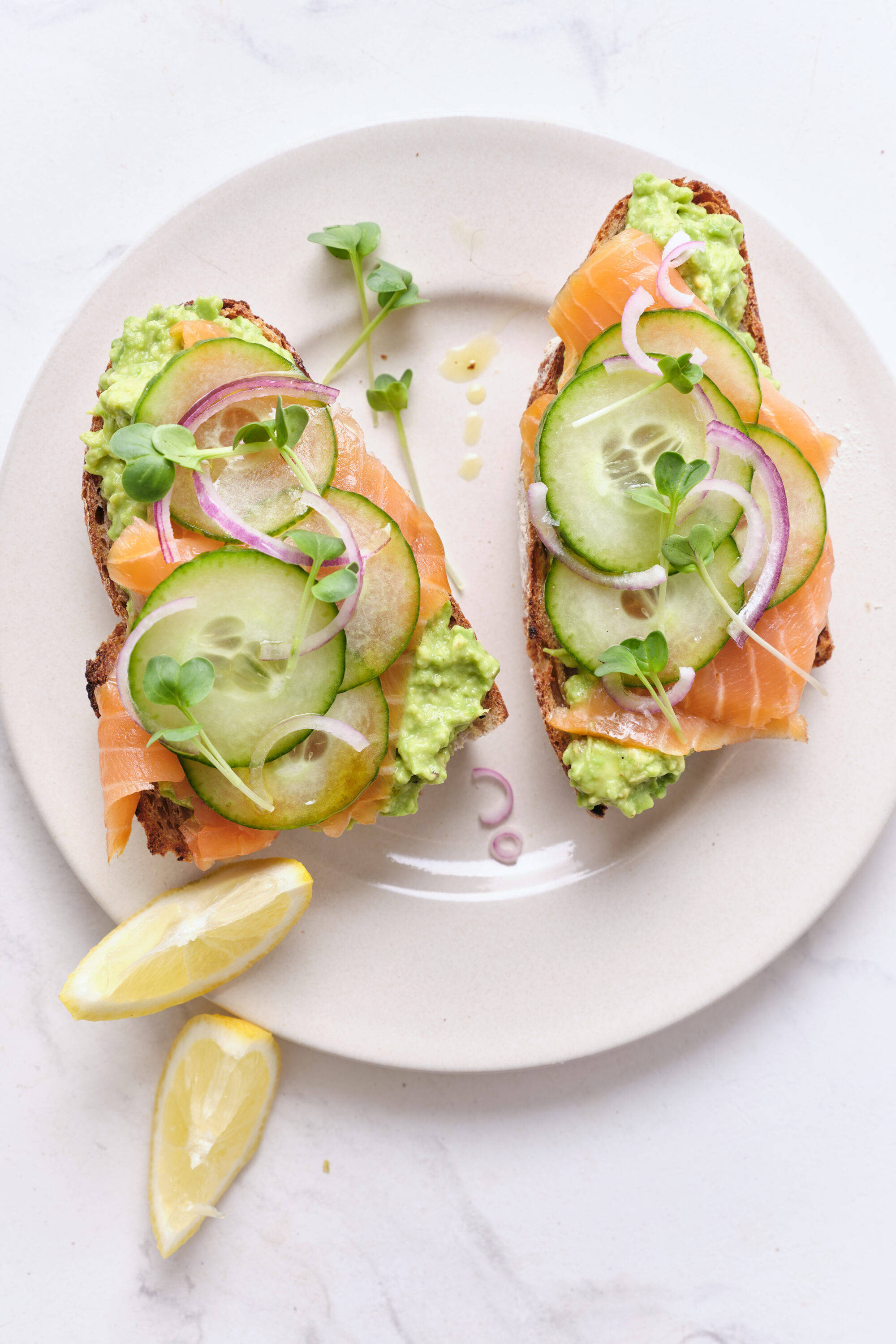Fresh smoked salmon avocado toast with cucumber, red onion, and microgreens on whole grain bread, served with lemon wedges. Healthy, protein-rich meal perfect for breakfast or brunch.