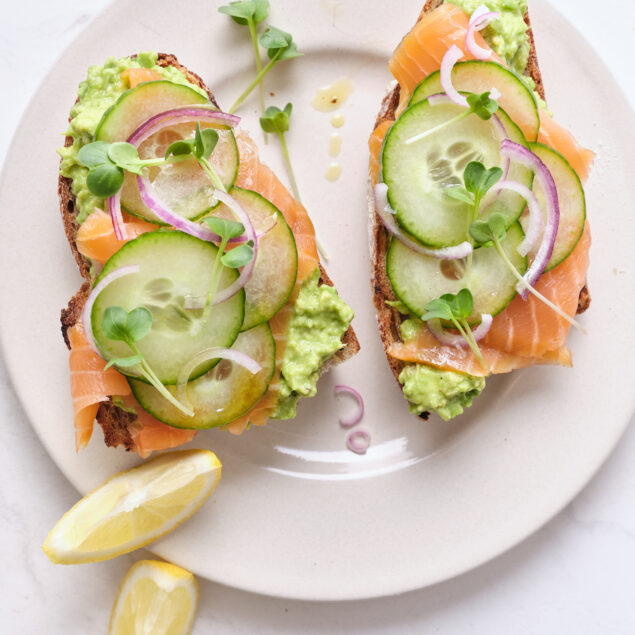 Fresh smoked salmon avocado toast with cucumber, red onion, and microgreens on whole grain bread, served with lemon wedges. Healthy, protein-rich meal perfect for breakfast or brunch.