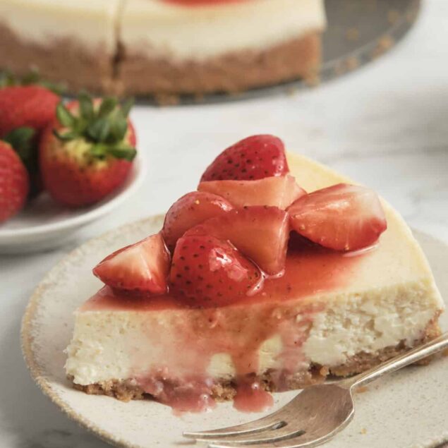 Fresh strawberry cheesecake topped with strawberry glaze, served on a white plate with whole strawberries and a sliced cheesecake in the background. Perfect for healthy dessert recipes on Food Faith Fitness.