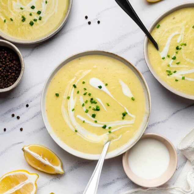 Creamy lemon soup with fresh chives, garnished with a swirl of cream, served in white bowls with lemon wedges and black peppercorns on a white wooden surface, highlighting healthy and flavorful meal options.