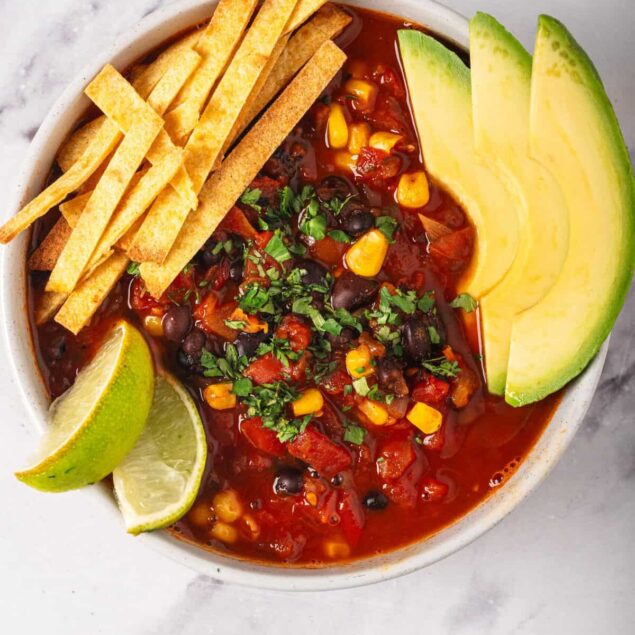 Savory vegan black bean chili served with fresh avocado slices, lime wedges, and crispy tortilla strips on a white plate, healthy plant-based food, gluten-free, high-protein, nutritious dinner option.