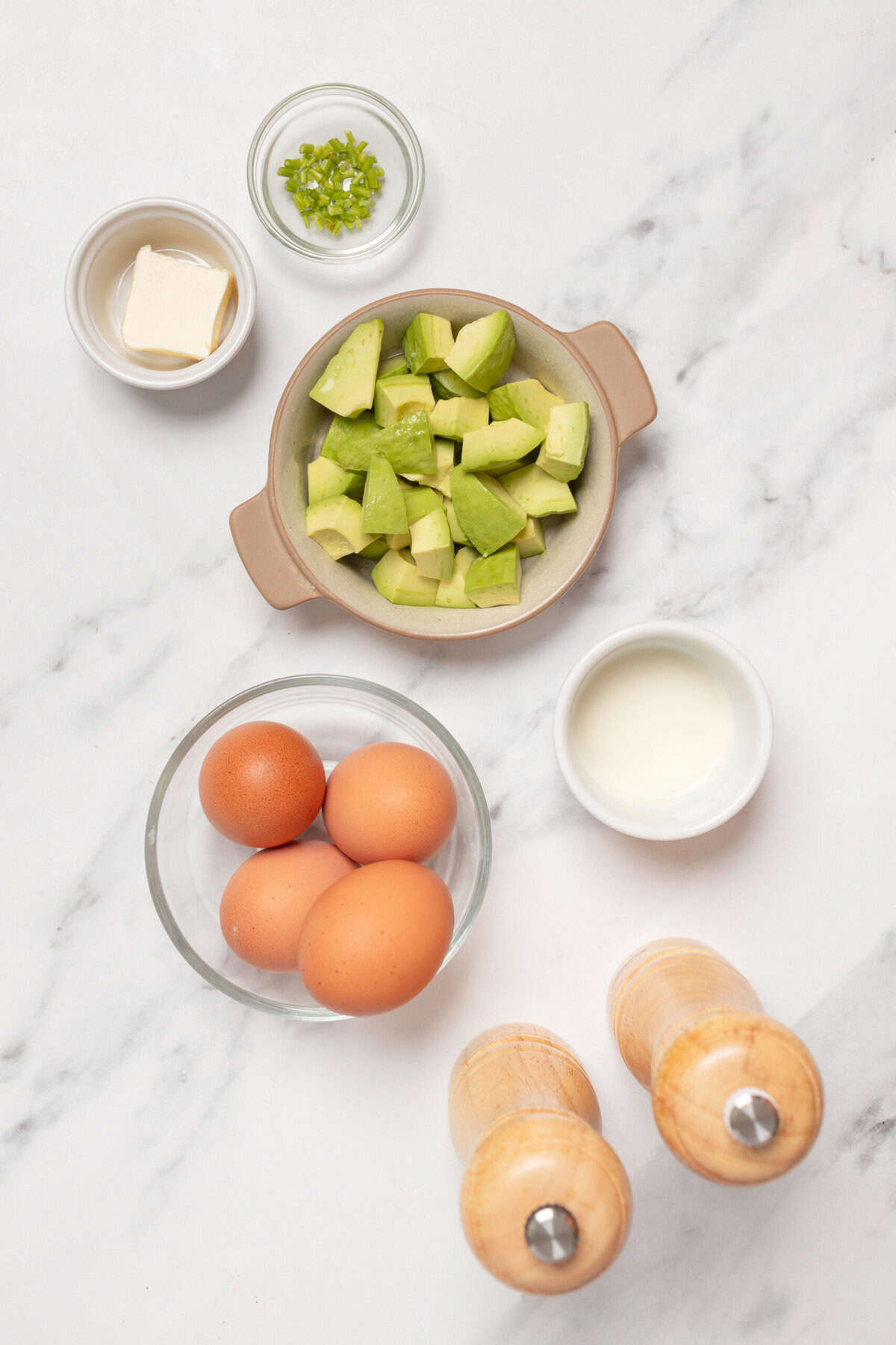 Fresh avocado slices, eggs, butter, chopped green onions, and seasonings on a white marble countertop, ideal ingredients for healthy breakfast or nutritious recipes.