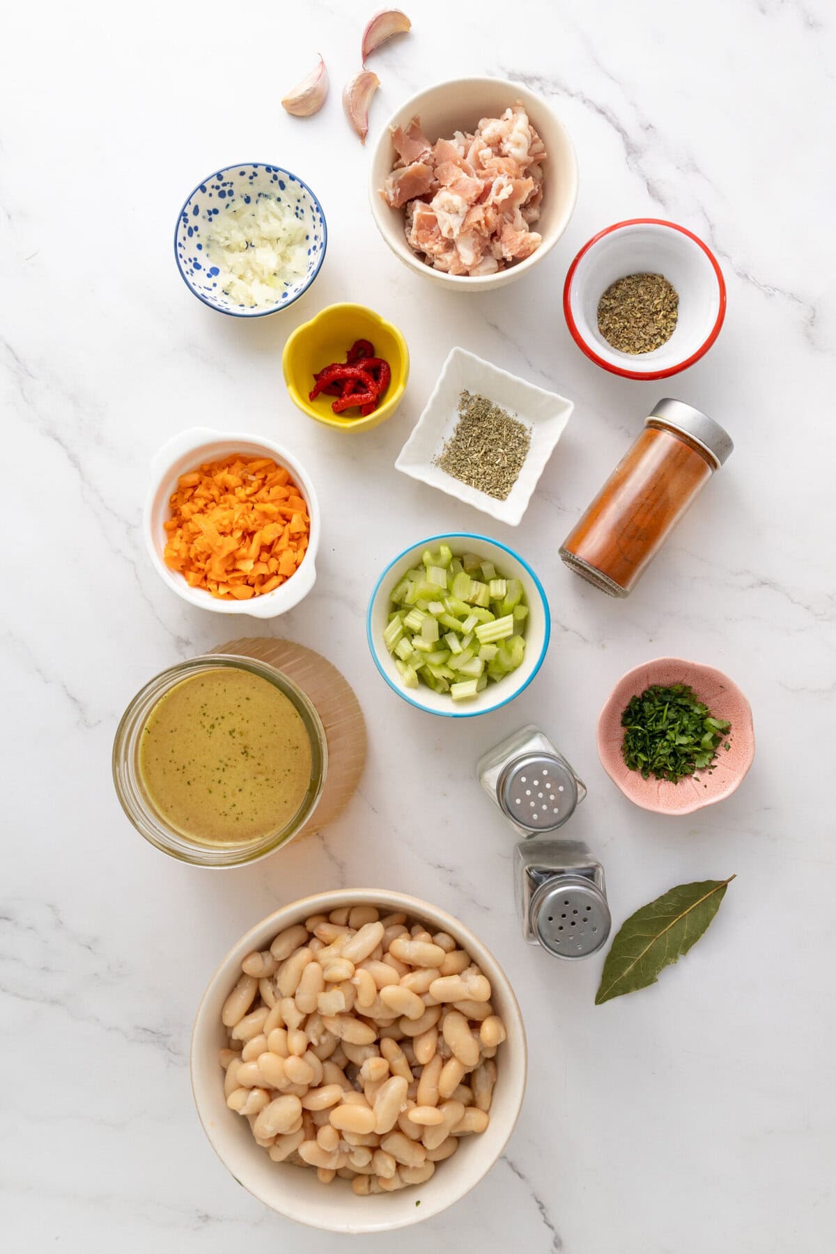 Sliced garlic, onions, carrots, celery, cooked cannellini beans, raw chopped chicken, spices, herbs, and vegetable broth arranged on a white marble surface for healthy meal preparation.