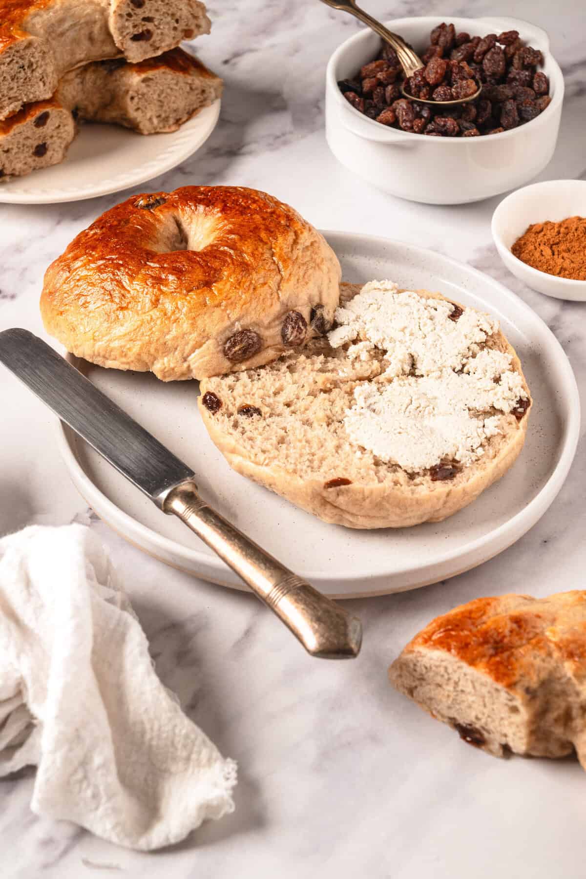 Cream cheese stuffed bagel with raisins on white plate, side of toasted bagel, dried fruit, spices, and bread on marble surface, healthy breakfast, high-protein snack, baking recipes, Food Faith Fitness.