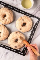 Soft, homemade chocolate chip bagels baking on a black cooling rack with a small bowl of melted butter. Perfect for a healthy breakfast or snack, these bagels combine wholesome ingredients with rich chocolate chips.