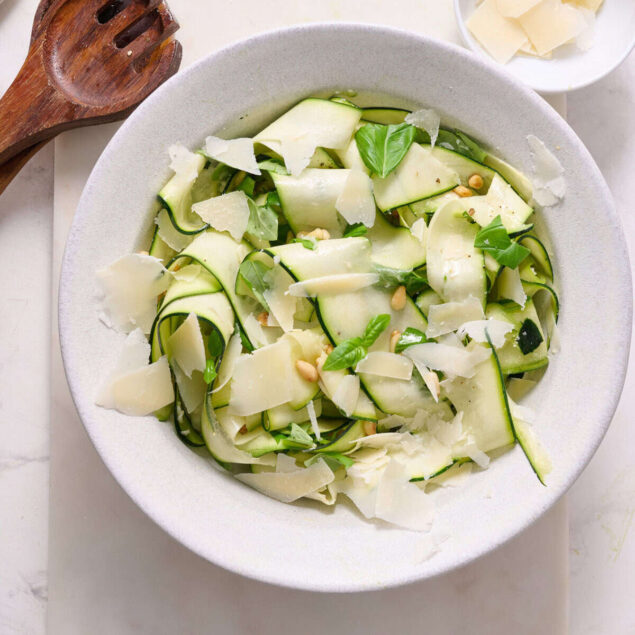 Cucumber salad with shaved Parmesan, fresh basil, pine nuts, and olive oil in a white bowl on a white surface.