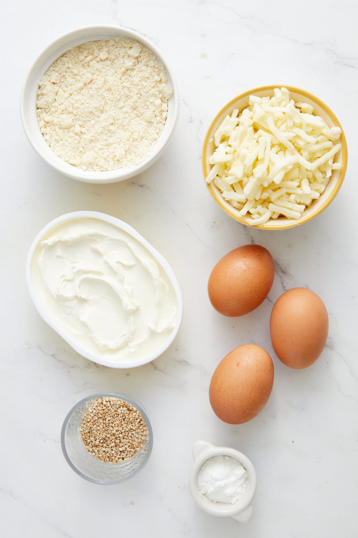 Cream cheese, shredded cheese, eggs, flour, sesame seeds, and sour cream on a white marble surface, ingredients for healthy recipes, nutritious cooking, and protein-rich meals.