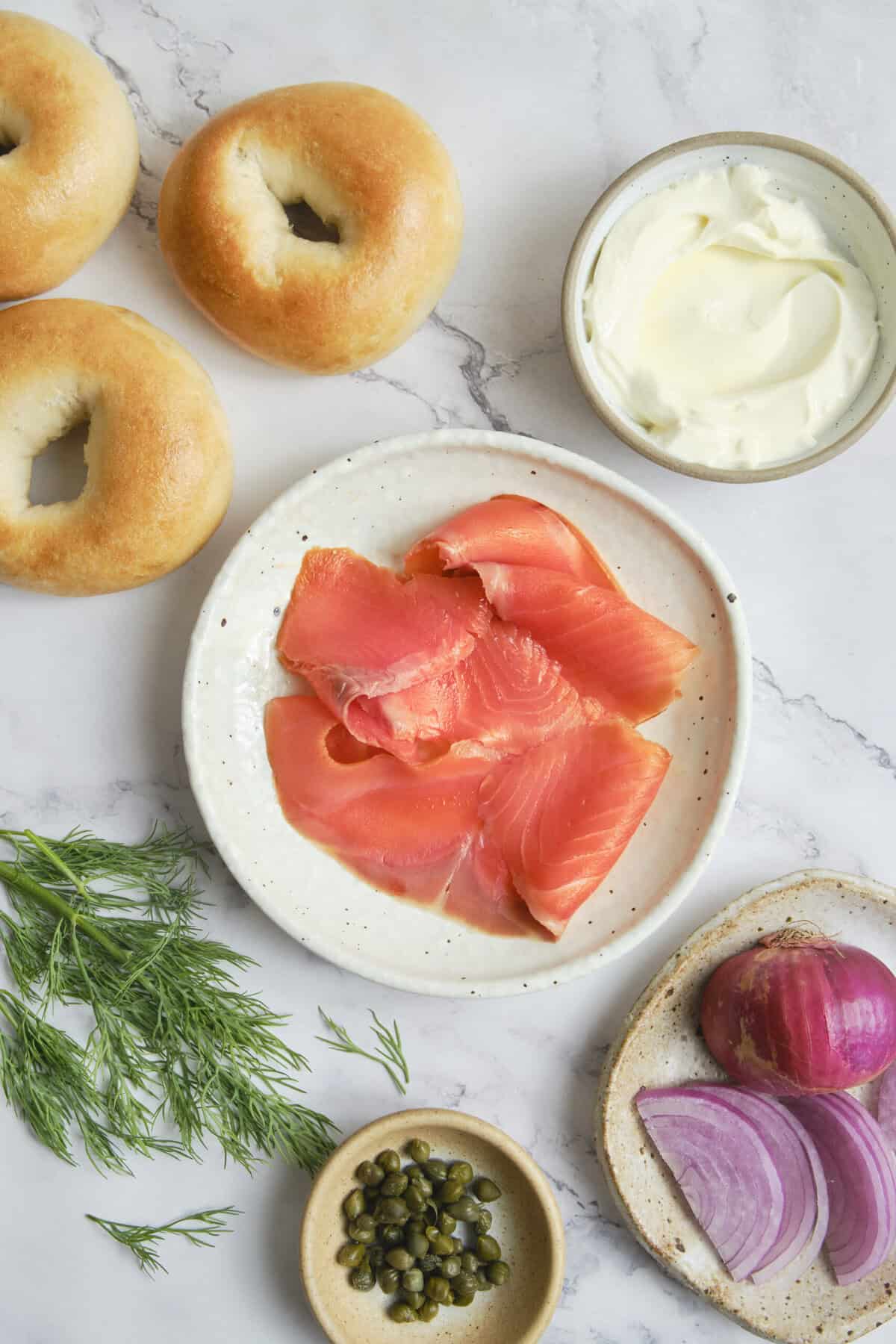 Fresh smoked salmon on a white speckled plate with ingredients like bagels, cream cheese, red onion, capers, and dill on a marble background for healthy, protein-rich breakfast ideas.