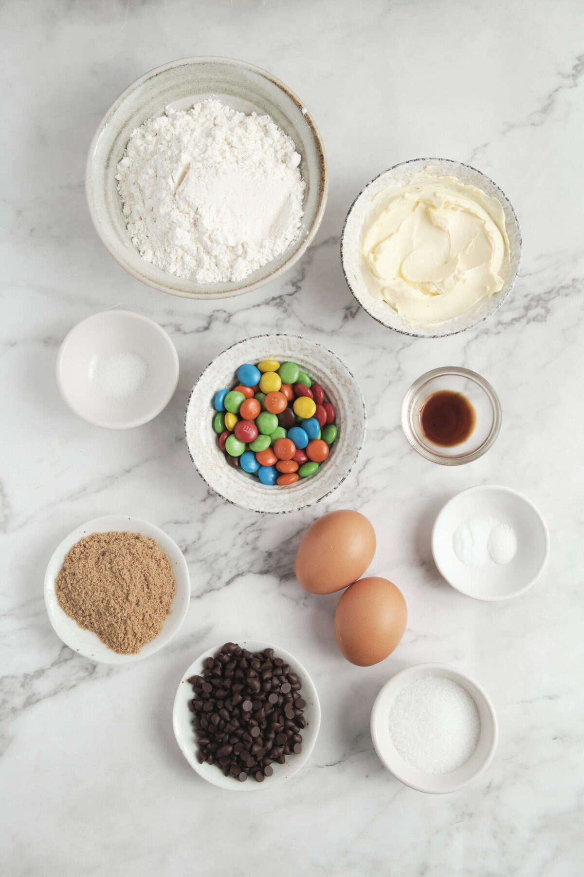 Colorful baking ingredients on marble surface, including chocolate chips, rainbow candies, eggs, flour, butter, vanilla extract, and cocoa powder for healthy dessert recipes.