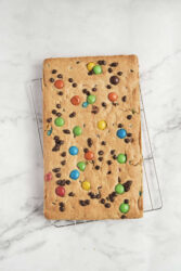 Colorful homemade chocolate chip cookie bar topped with M&Ms cooling on a wire rack on a white marble surface. Perfect for healthy snacks and delicious desserts in food, fitness, and wellness recipes.