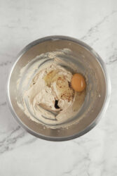 Cream cheese mixture with egg in a stainless steel bowl, ready for baking or cooking, on a white marble surface.
