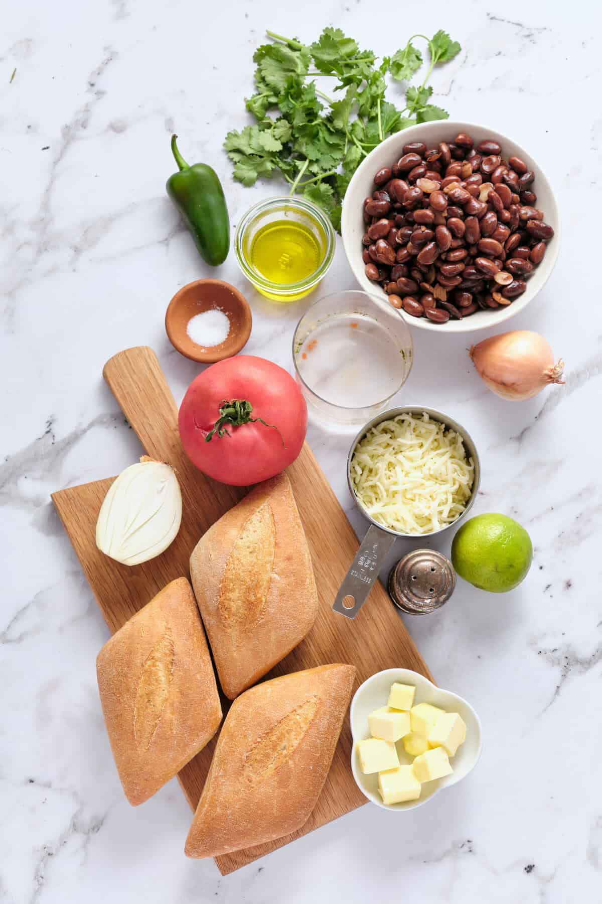Fresh ingredients for black bean cheese hoagies including bread, cheese, black beans, tomato, onion, lime, jalapeno, cilantro, olive oil, and seasonings on a marble surface.
