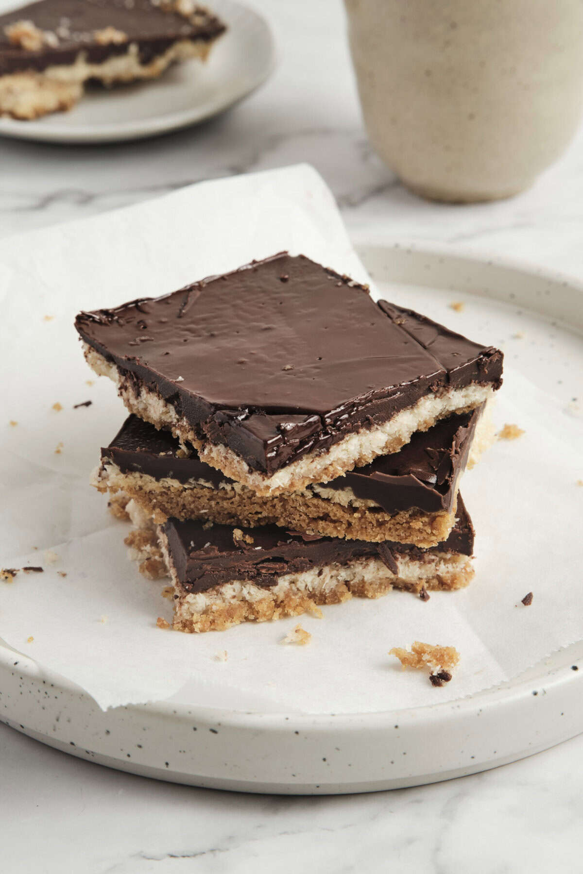 Decadent layered chocolate peanut butter bars on white plate with a blurred background of additional treats and cups. Perfect for healthy dessert recipes and indulgent snack ideas from Food Faith Fitness.