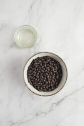 Rich dark chocolate chips in a white ceramic bowl with a glass of water on white marble surface, healthy dessert ingredients, baking, chocolate recipes, food photography.
