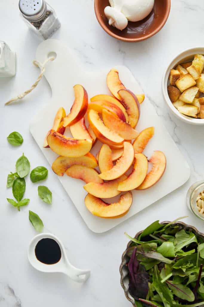 Fresh sliced peaches on a cutting board with basil leaves, salad greens, croutons, balsamic vinegar, garlic, and seasonings, showcasing healthy eating and meal prep ideas from Food Faith Fitness.
