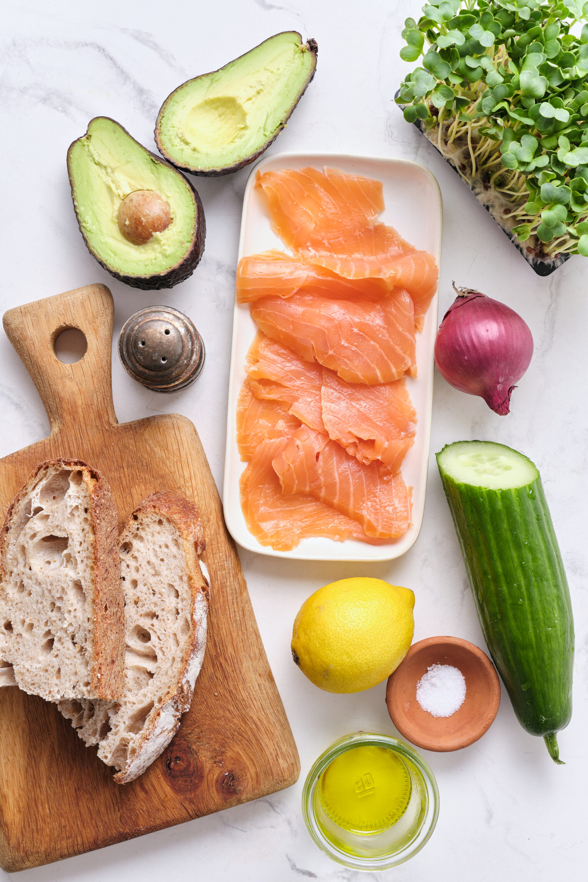 Fresh salmon slices, avocado, cucumber, red onion, microgreens, whole grain bread, lemon, olive oil, salt, and pepper on a white marble surface, perfect for healthy eating and nutritious meal ideas.