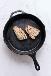 Crusty sourdough bread slices in a black cast iron skillet on white marble background for healthy eating and nutrition.