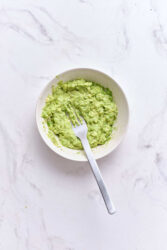 Creamy avocado mash in a white bowl with a fork on a light marble surface. Healthy, nutritious, and perfect for snacks or toppings. Great for meal prep and weight loss.