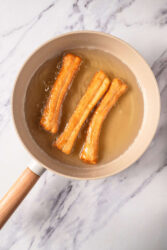 Cocoa churros frying in a beige frying pan with hot oil on a white marble countertop.